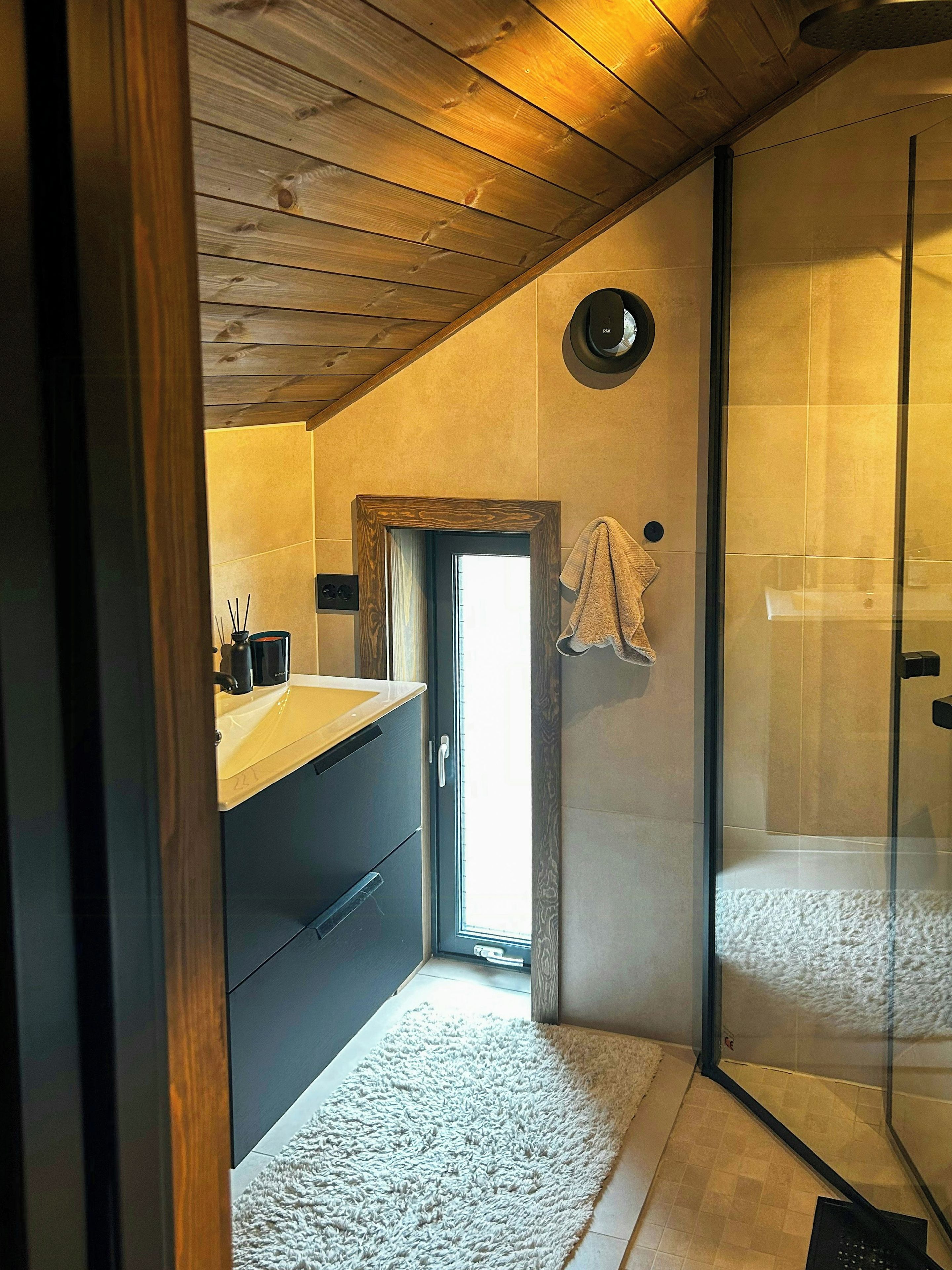Een badkamer met een houten plafond, een glazen douchecabine, een wastafel en een handdoek aan de muur, plus een klein raam.