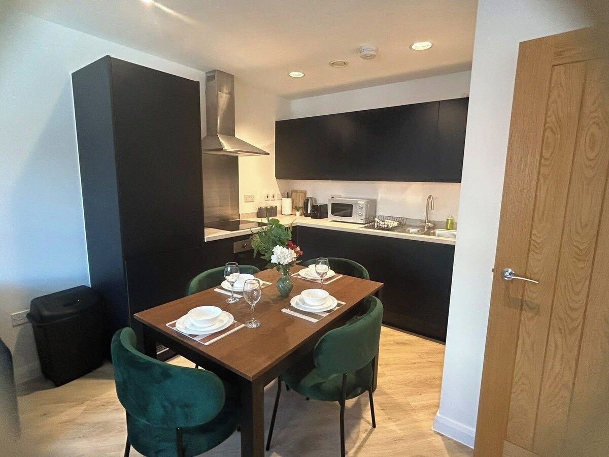 A dining area with a wooden table set for four, green chairs, and a kitchen in the background.