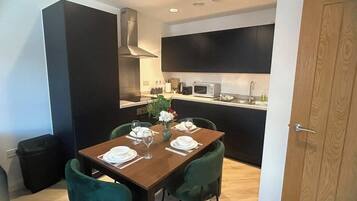 A dining area with a wooden table set for four, green chairs, and a kitchen in the background.
