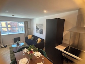 A modern living room with a dining area, a sofa, a painting on the wall, and a kitchen with a stove. - 1BR Flat in Andover | Near Amenities & Transports (Hampshire)