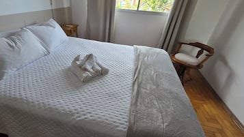 A neatly made bed with white linens and a folded towel, a wooden nightstand with a lamp, and a chair with a cushion.