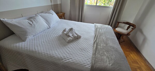A neatly made bed with white linens and a folded towel, a wooden nightstand with a lamp, and a chair with a cushion.
