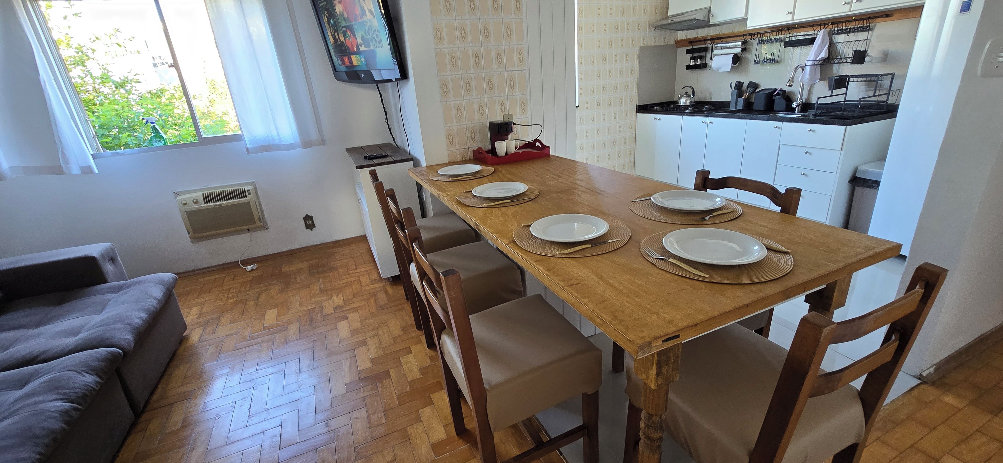 Un comedor con una mesa de madera preparada para cuatro, rodeada de sillas, en una habitación con suelo de diseño espiga y un sofá gris.