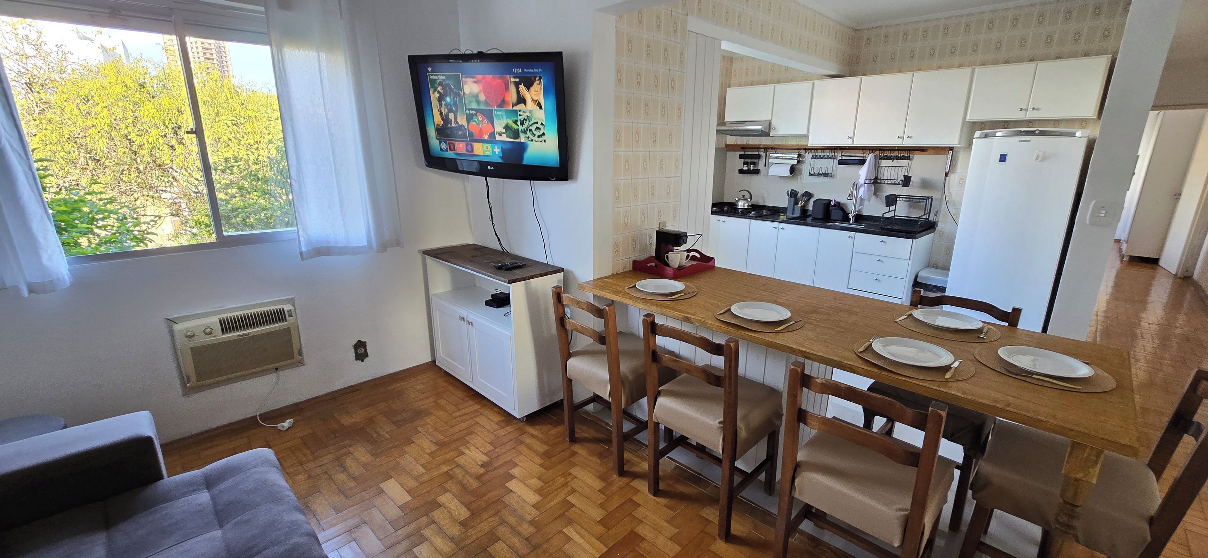 Kompakti keittiö, jossa on valkoiset kaapit, ruokapöytä, jonka päällä on lautasia ja aterimia, seinälle asennettu televisio ja ikkuna, josta avautuu näkymä puita kohti.