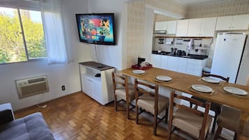 Kompakti keittiö, jossa on valkoiset kaapit, ruokapöytä, jonka päällä on lautasia ja aterimia, seinälle asennettu televisio ja ikkuna, josta avautuu näkymä puita kohti.