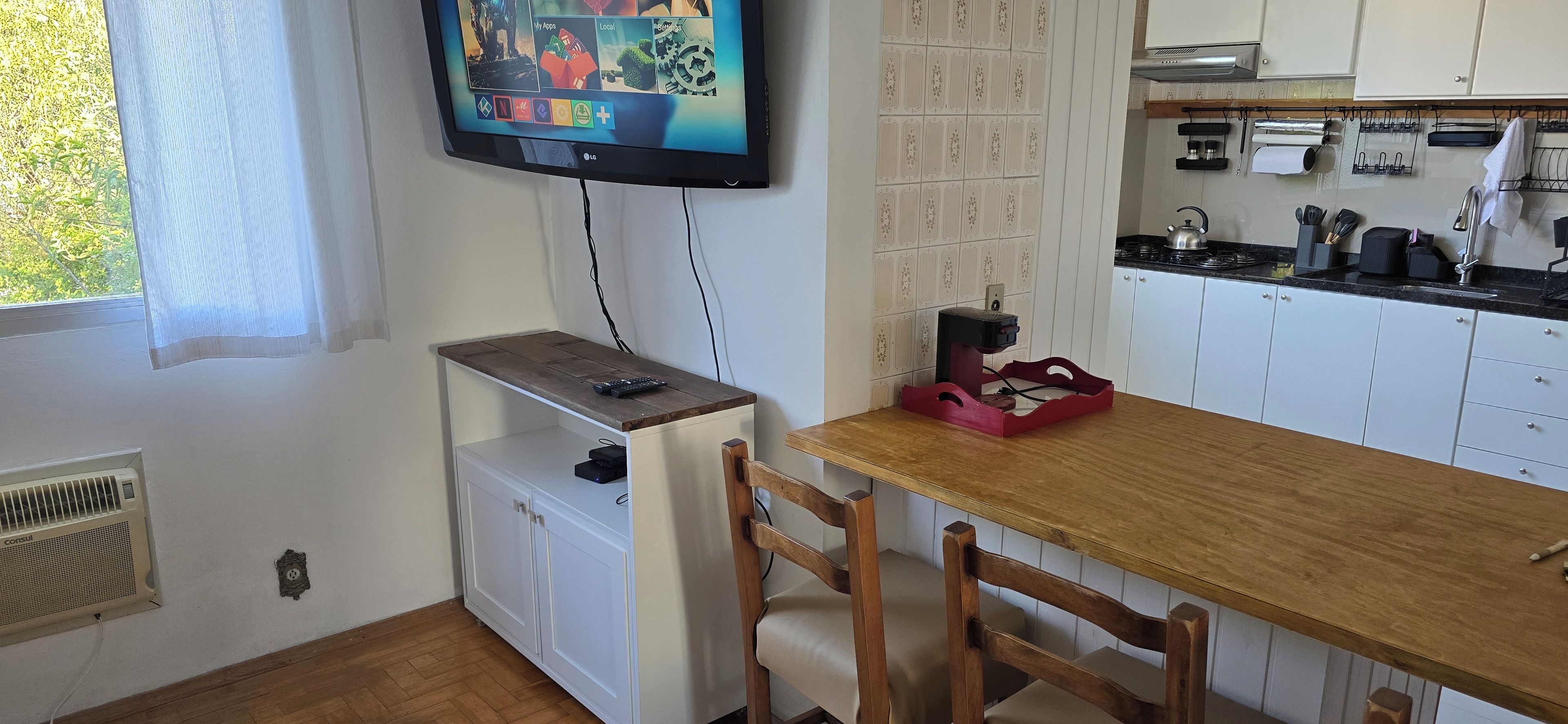 Een keuken met een houten tafel, stoelen, een aan de muur bevestigde televisie en een raam met gordijn.