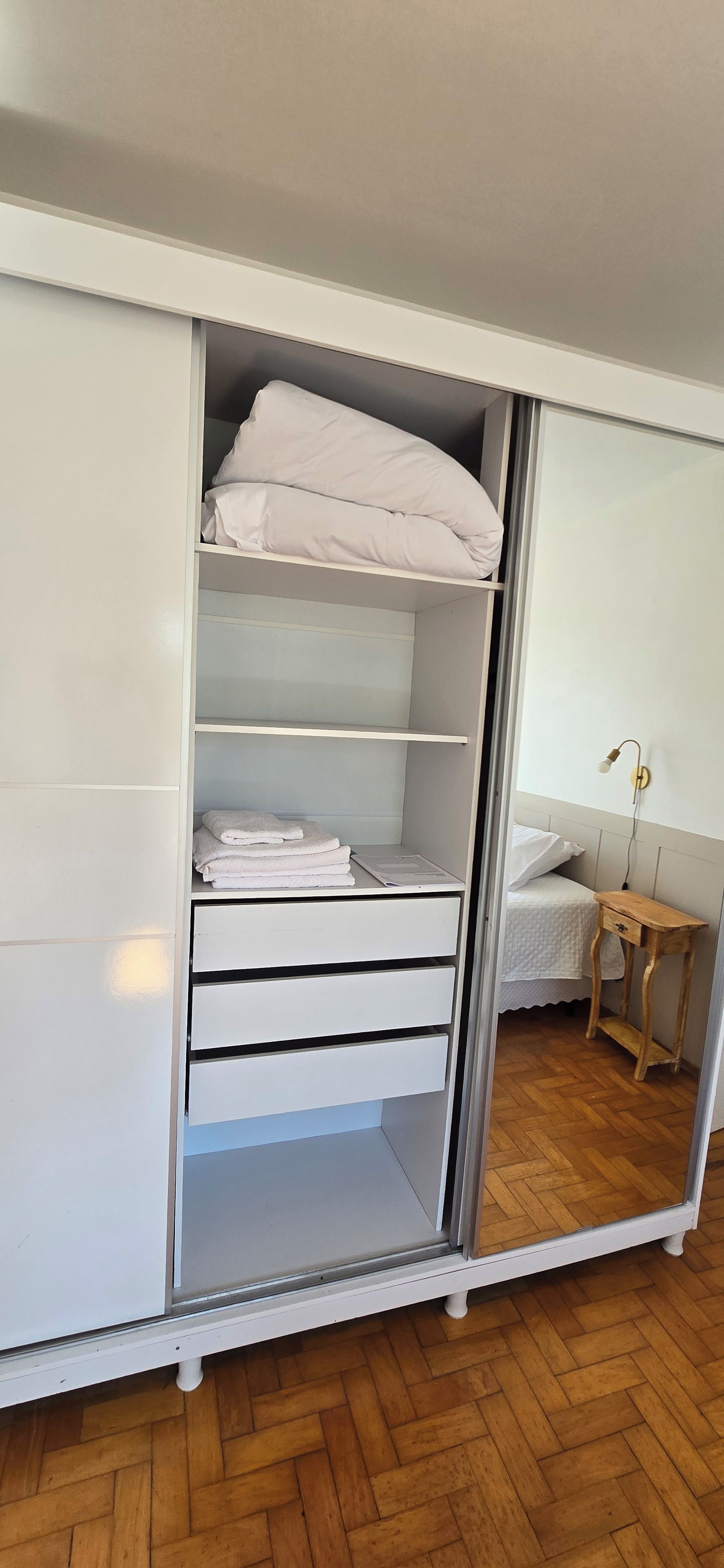 A white wardrobe with multiple shelves and drawers, a wooden bedside table, and a bed with white bedding.