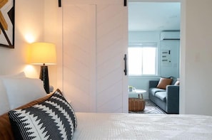 A bedroom with a bed, a nightstand, a lamp, and a view into a living area with a sofa and a window. - Melrose 1BR Getaway with Pool  by Barcle (Nashville)