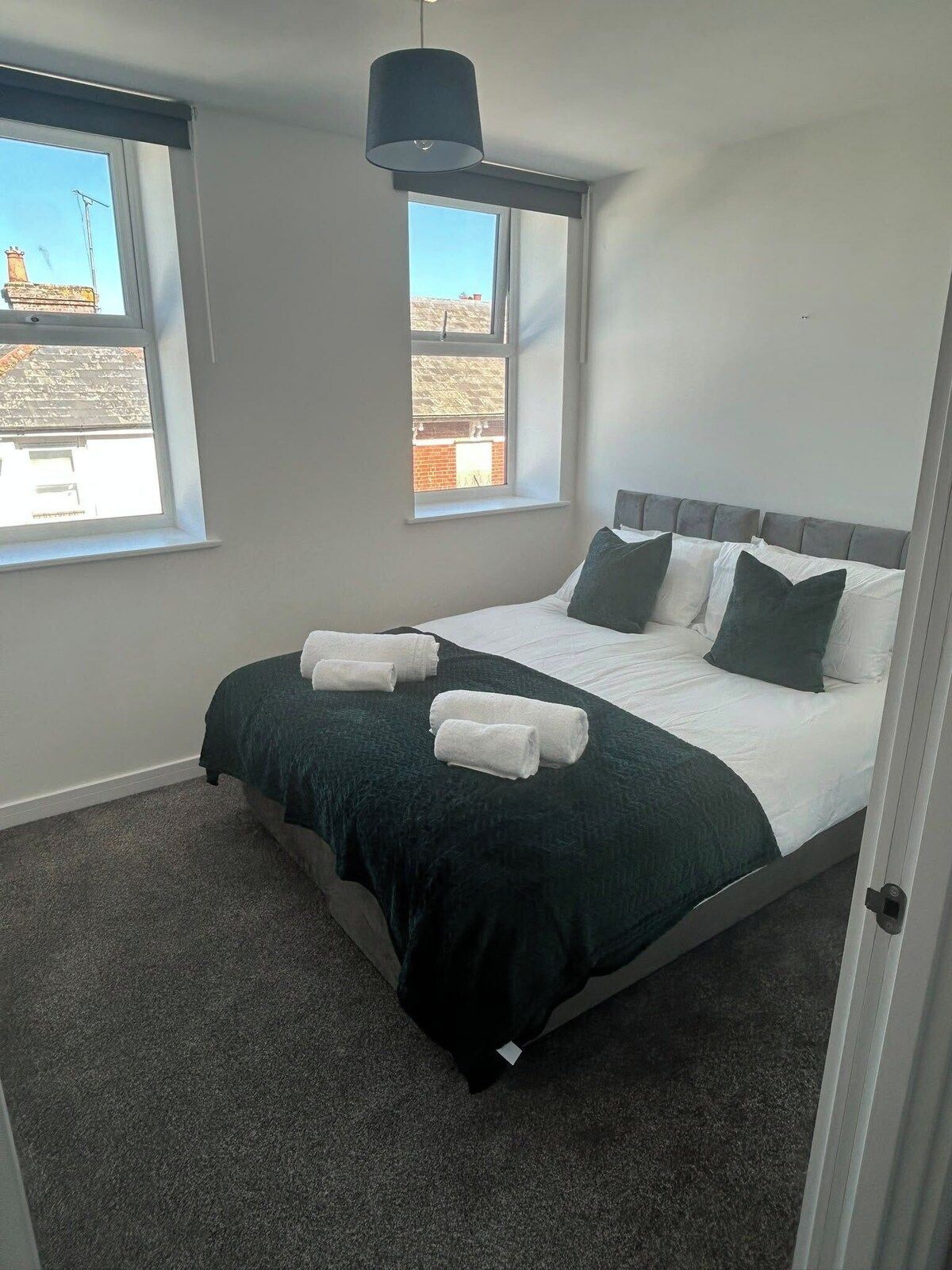 Une chambre à coucher avec un lit, deux fenêtres, un luminaire suspendu et une vue sur l’extérieur.