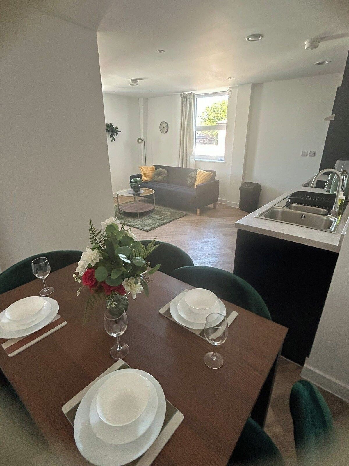 A dining area with a table set for four, a sofa in the background, and a kitchen area with a sink.