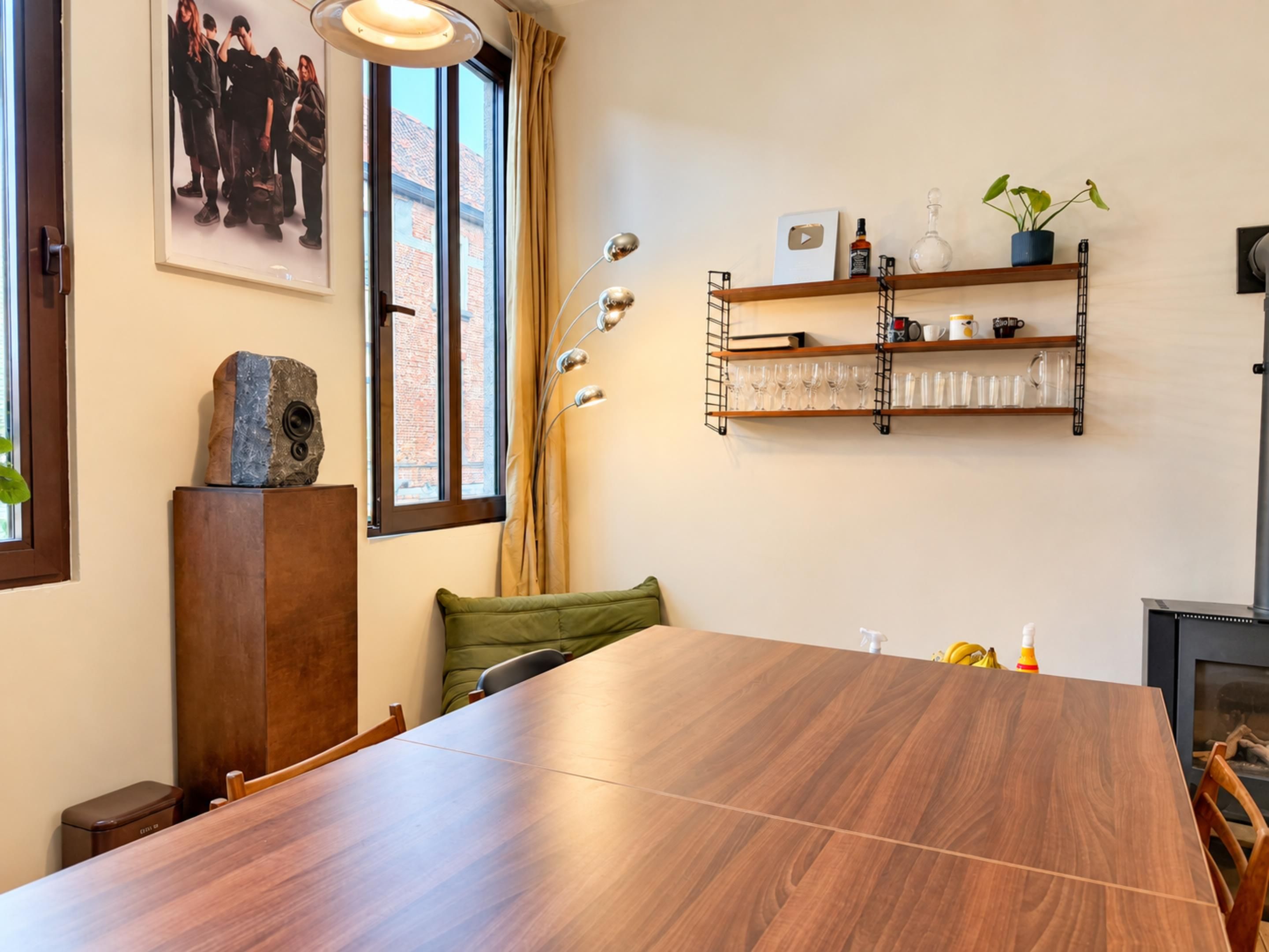 Uma sala de jantar com uma mesa de madeira comprida, cadeiras, um travesseiro verde, uma luminária pendente, uma estante na parede com copos e um quadro emoldurado.