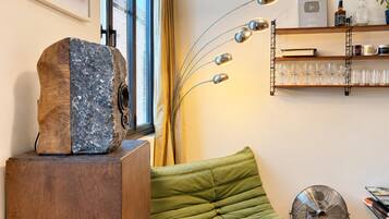 Quarto com uma escultura de madeira em pedestal, um banco estofado verde, um ventilador de chão e uma estante com livros e copos.