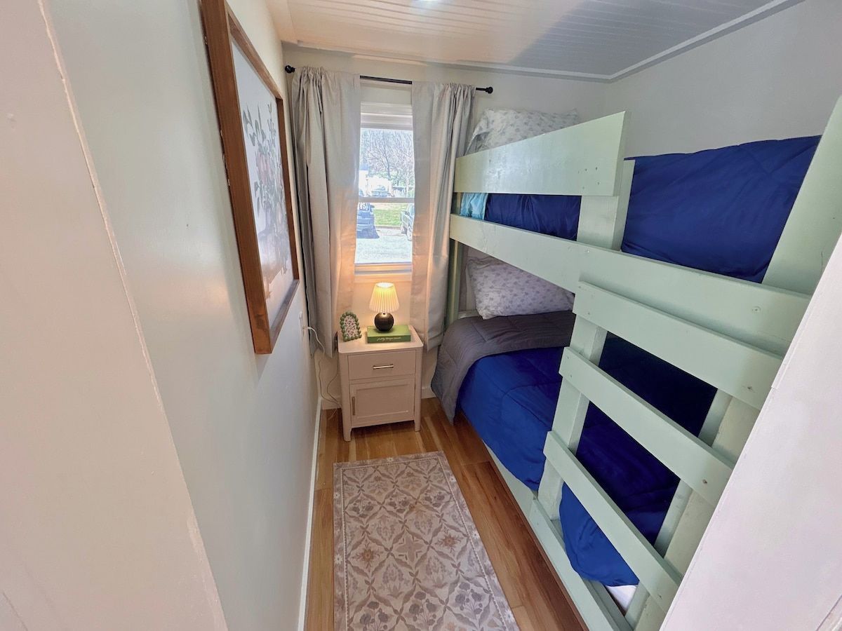 A narrow room with bunk beds, a bedside table with a lamp, and a patterned rug.