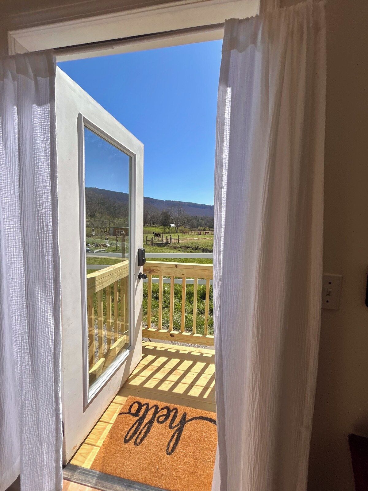 An open door to a balcony with a wooden railing, a doormat with the word 'home,' and a view of a green landscape with trees and hills.