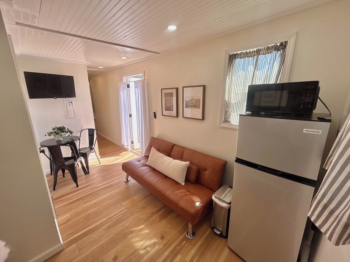 A compact living space with a sofa, a dining table, a TV mounted on the wall, and a refrigerator.