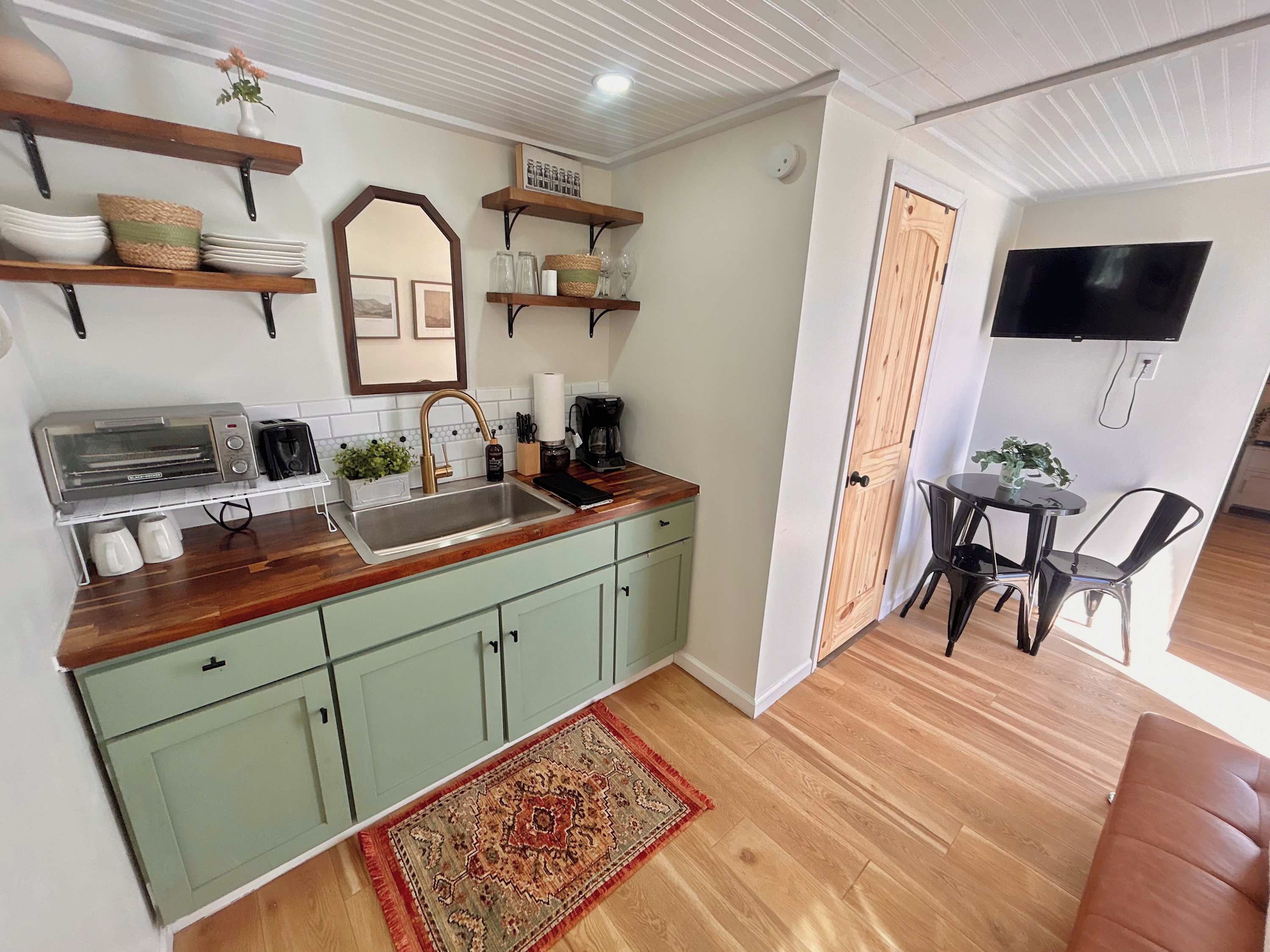 A compact kitchen with a built-in toaster, a sink, and a coffee maker. There is a dining area with a table and chairs, and a TV mounted on the wall.
