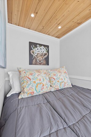 Une chambre avec un plafond en bois, une couvre-lit grise, un coussin à motifs floraux colorés et un tableau représentant une vache accroché au mur.