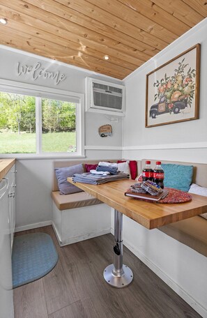 A corner room with a wooden ceiling, a window with a view of greenery, a wall-mounted air conditioner, a framed artwork, a bench with towels, and a small table with snacks. - Quail Run Farm- Rustic Tiny Home 1 (Chattanooga)