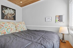 Une chambre avec une couvre-lit grise, un coussin à motifs floraux, une lampe fixée au mur et un tableau représentant une vache entourée de fleurs.