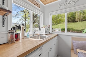 Une cuisine dotée d’un plan de travail en bois, de meubles blancs, d’un évier et offrant une vue sur un paysage verdoyant grâce à de grandes fenêtres.