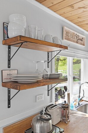 A kitchen with wooden shelves holding glassware and plates, a sign saying 'BE OUR Guest,' and a view of the outdoors. - Quail Run Farm- Rustic Tiny Home 1 (Chattanooga)