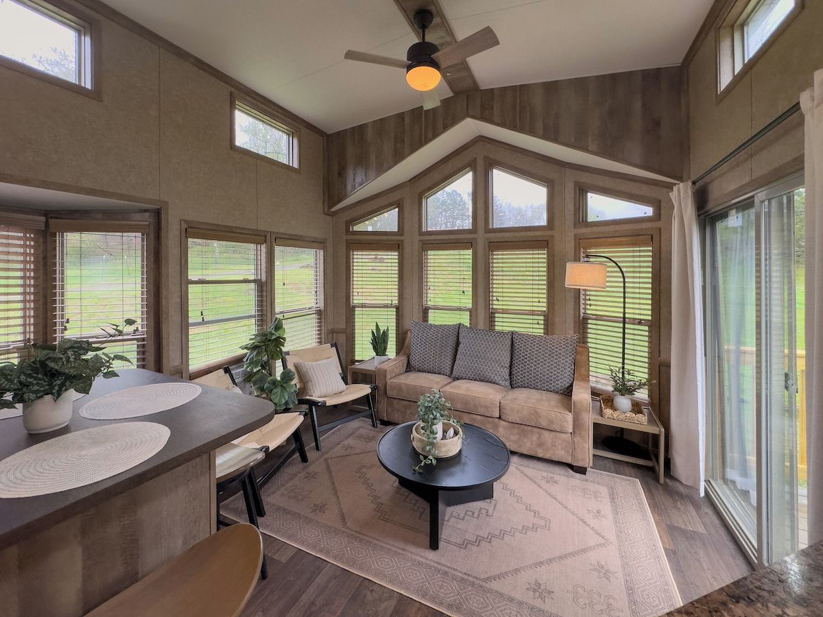 A spacious room with a high ceiling, large windows, a ceiling fan, and a cozy seating area with a sofa and chairs.