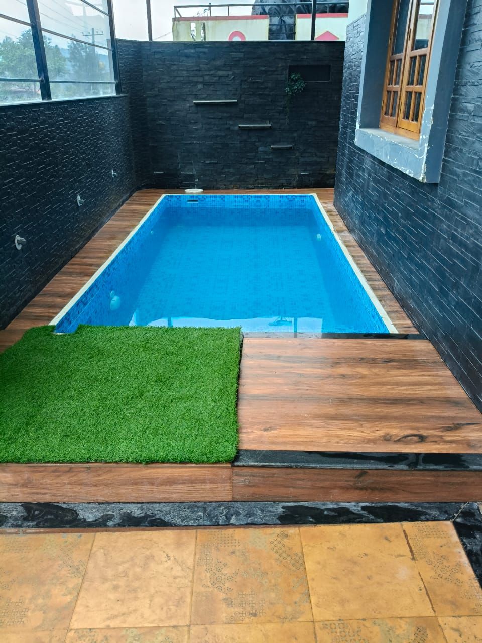 Una piscina rectangular con un deck de madera y césped artificial en una esquina.
