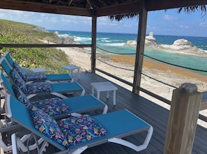 A wooden deck with lounge chairs and a small table, overlooking a beach with clear blue water and rocky outcrops. - Bahama Dreamin' Ocean Front Villa 1/1 (Long Island)