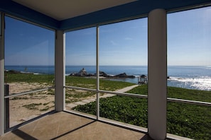 A room with large windows overlooking a beach with rocks and greenery.