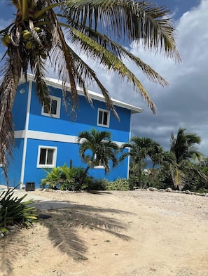A two-story building with a blue exterior and white trim, surrounded by palm trees and a sandy ground. - Bahama Dreamin' Upstairs Oceanfront Villa 2/2 (Long Island)
