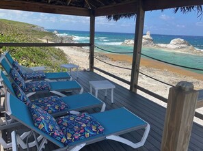 A wooden deck with lounge chairs and a small table, overlooking a beach with clear blue water and rocky outcrops.