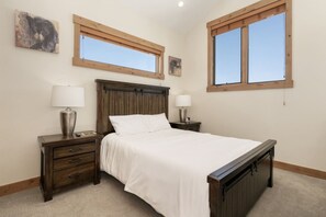 A bedroom with a wooden bed, two bedside tables with lamps, a nightstand, and two windows. - Mountain Escape With Hot Tub & Fire Pit (Fraser)
