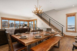 A spacious dining area with a wooden table set for six, a staircase, and a large window offering a mountain view. - Mountain Escape With Hot Tub & Fire Pit (Fraser)