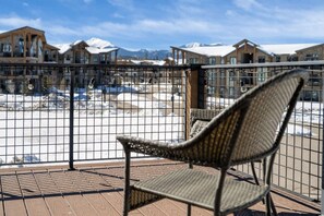 A balcony with a wicker chair, a metal fence, and a view of snow-covered mountains and buildings. - Mountain Escape With Hot Tub & Fire Pit (Fraser)