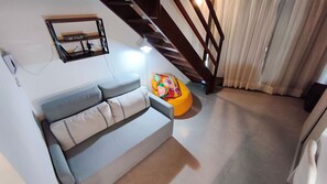 A living room with a grey sofa, a colorful ottoman, a staircase, and a window with curtains. - Carneiros Atlantico Beira-mar - Proximo da Igrejinha (Tamandaré)