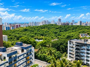 A high-rise building with multiple floors and balconies, surrounded by dense green foliage and a clear blue sky. - Studio Vista Mar  (Salvador)