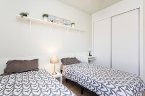A bedroom with two beds, a bedside table with a lamp, a shelf with plants and photos, and a wardrobe. - Home2Book Bright Abades Apt, Pool & Gym (Abades)