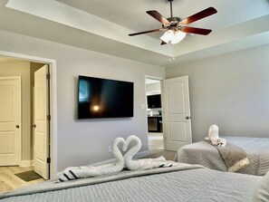 A hotel room with a bed, a television mounted on the wall, a ceiling fan, and a door leading to another room. - The Cozy Pond House — Spacious, Cozy, Peaceful (Olive Branch)