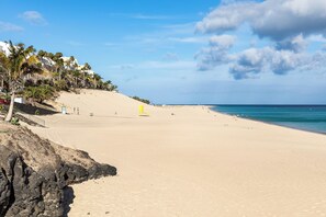 A wide sandy beach with a clear sky, palm trees, and a rocky cliff. - Home2Book Morro Jable Coastal Studio by the Beach (Solana Matorral)