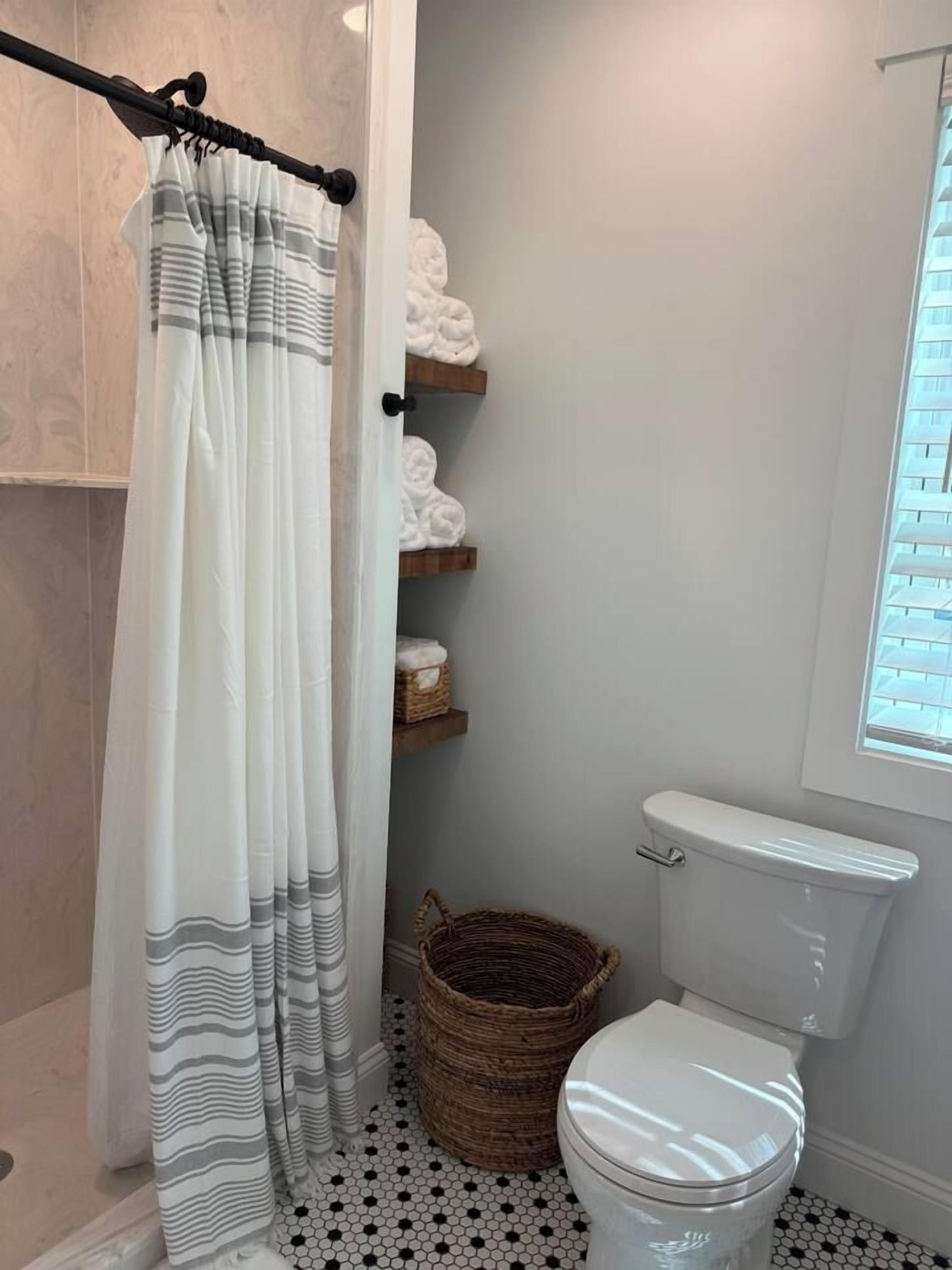 Une salle de bain avec un rideau de douche, des toilettes, un panier en osier et des étagères avec des serviettes.