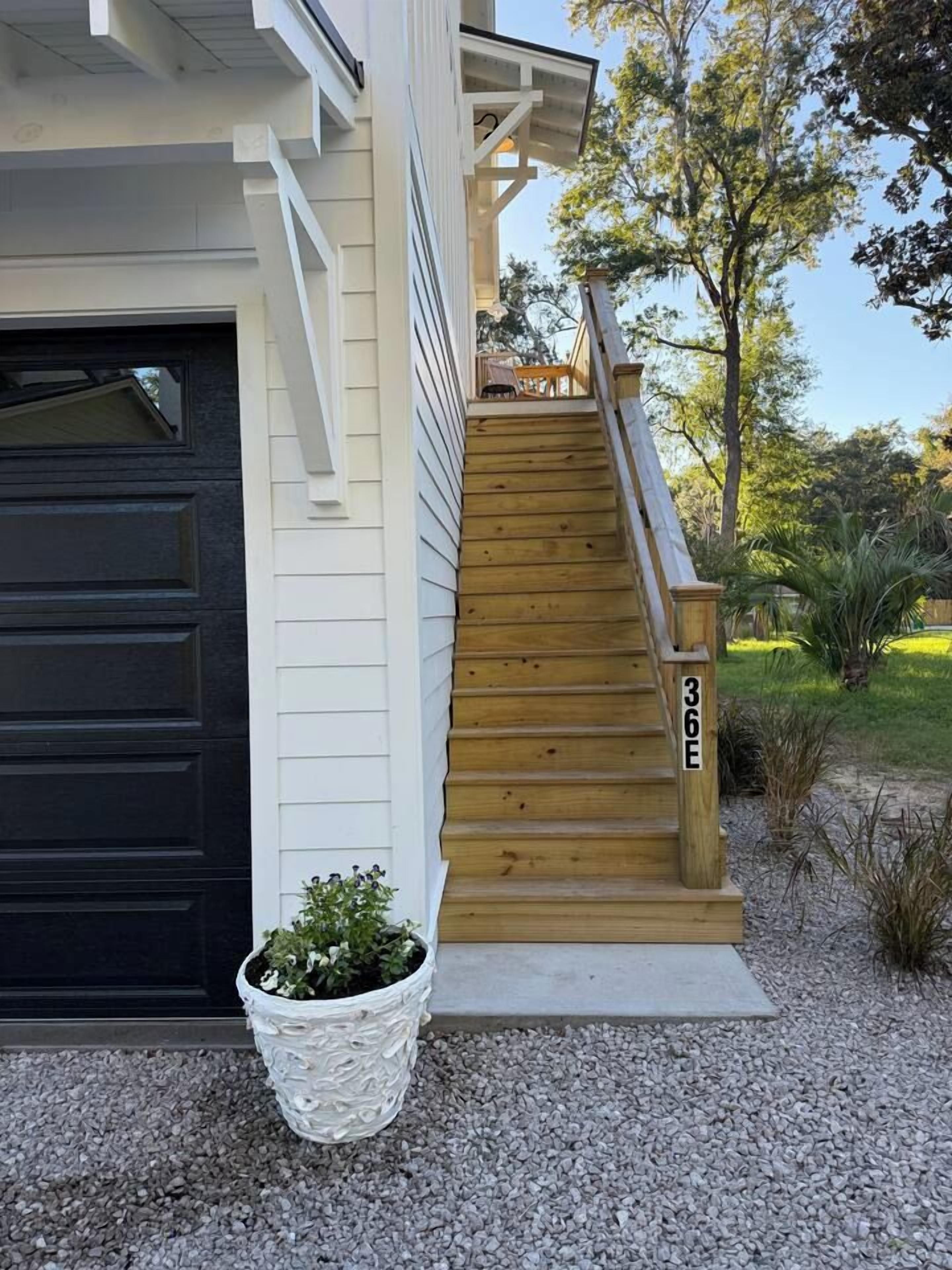A residential house with a wooden staircase leading to a deck, a black garage door, and a potted plant in the foreground.