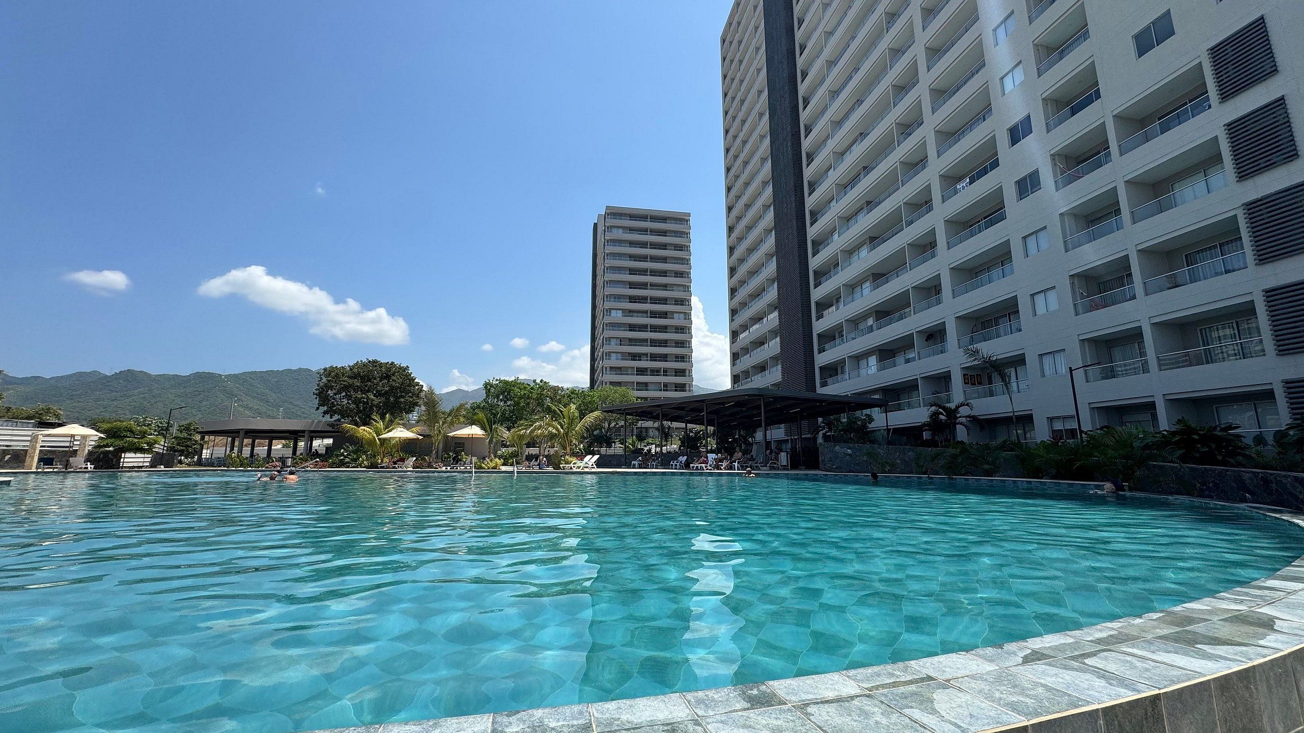 Una piscina amb aigua cristal·lina, envoltada d'una zona coberta amb gandules, i edificis alts al fons.