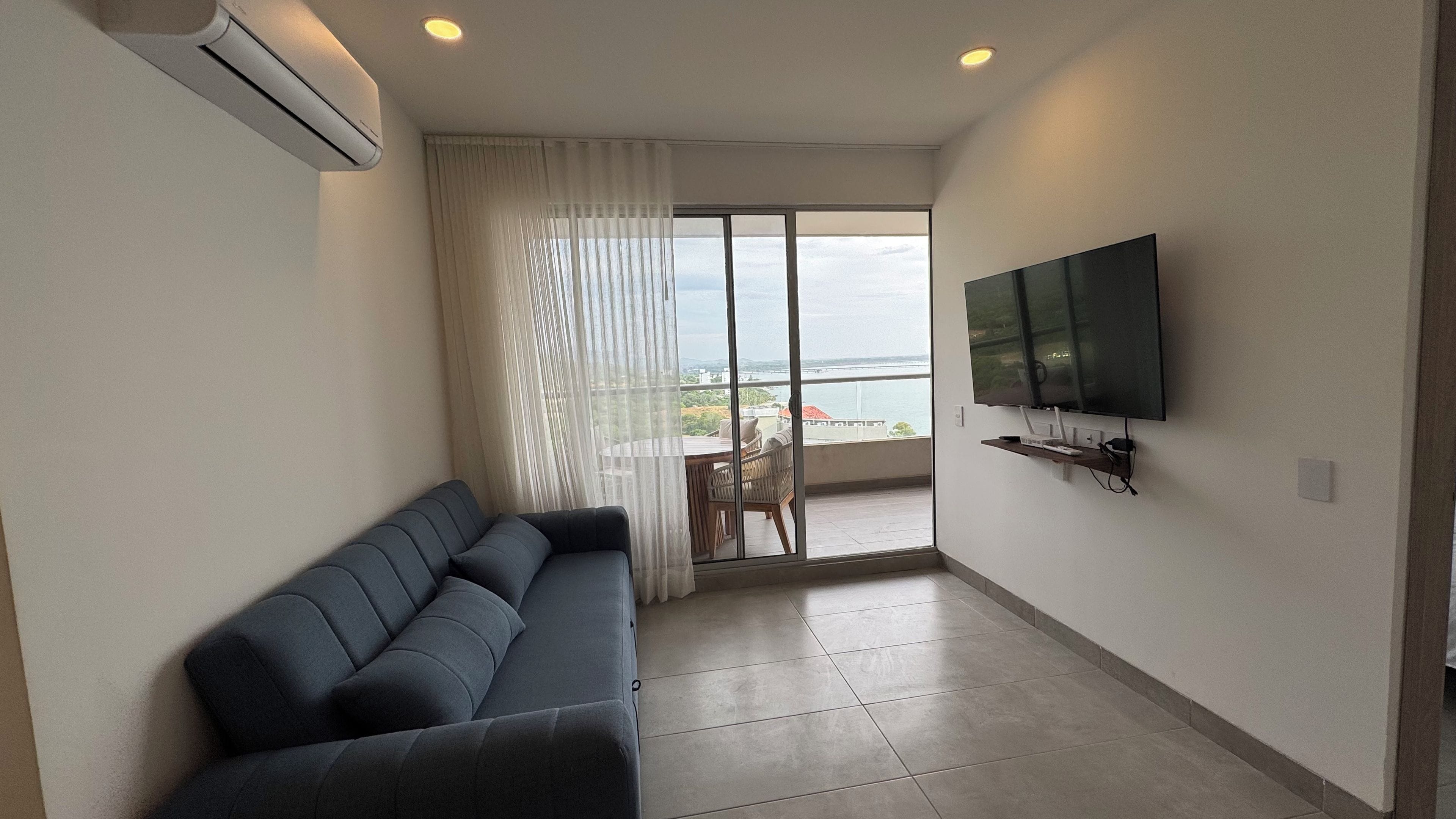 Una sala moderna con un sofá, un televisor de pantalla plana montado en la pared y un ventanal corredizo que da a un balcón con vistas.