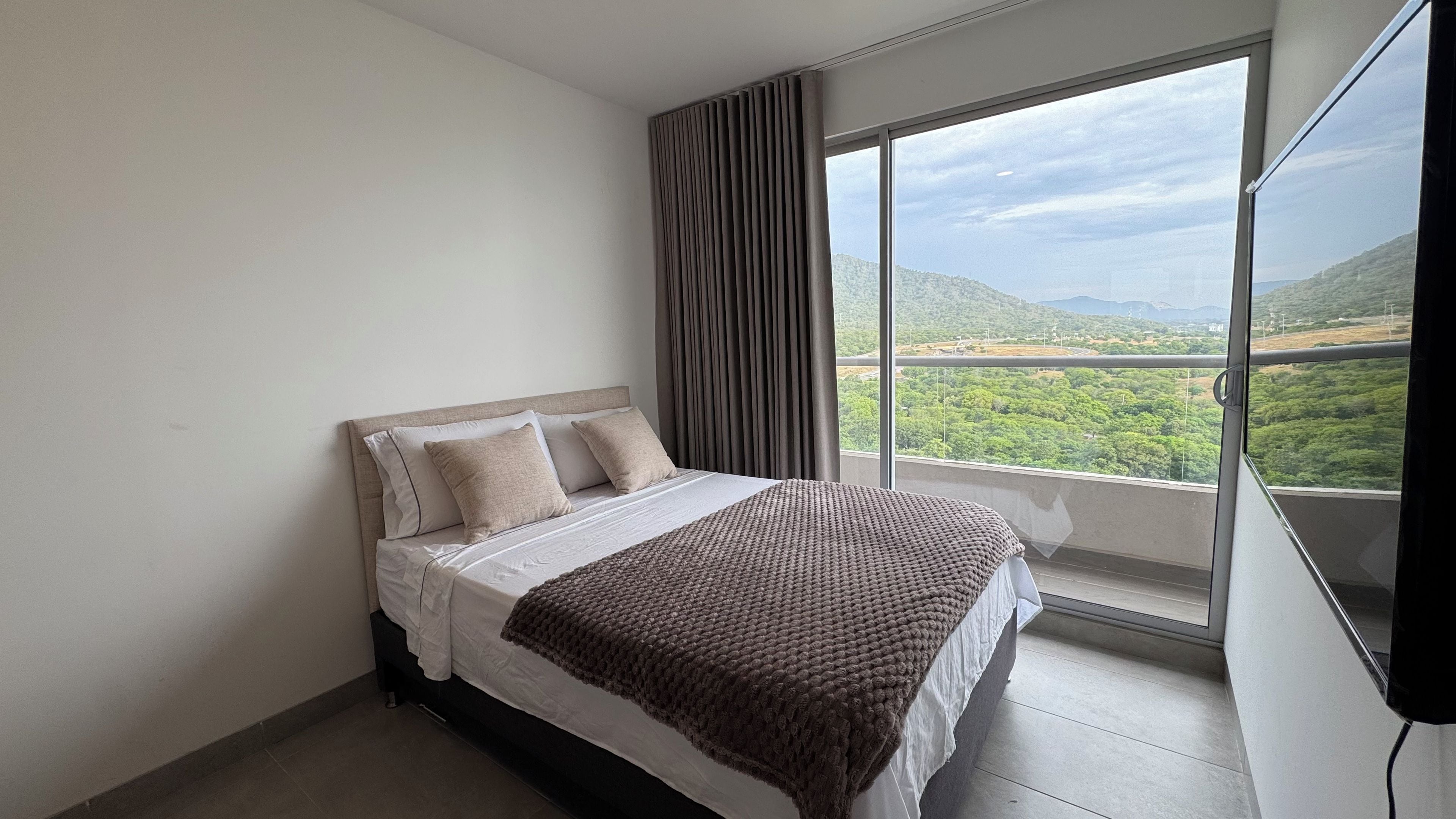 Un dormitorio con una cama grande, vista a un paisaje verde a través de una ventana y una cortina.