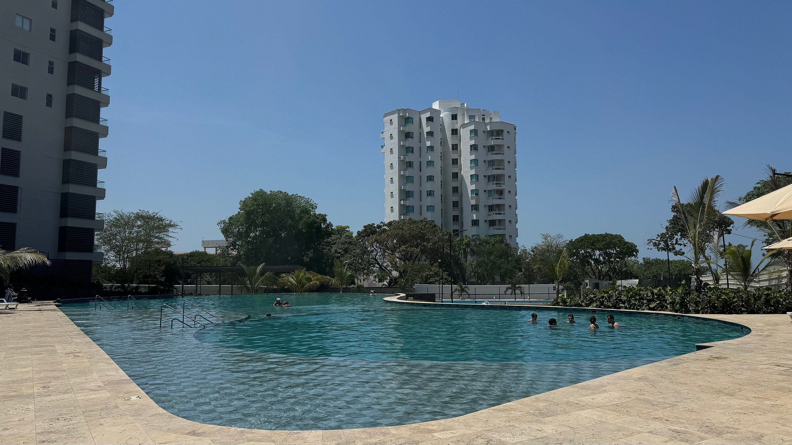 Piscina com pessoas na água, cercada por árvores e um prédio alto ao fundo.