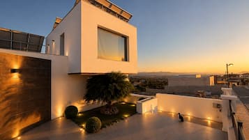Uma casa moderna de dois andares com telhado plano, janelas amplas e área externa bem iluminada, com um pequeno jardim e um gato.