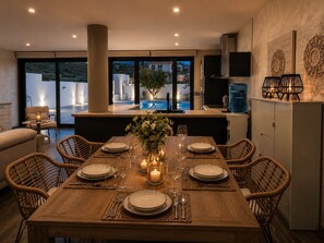 A dining area with a wooden table set for six, surrounded by wicker chairs, with a view of a swimming pool and a kitchen in the background. - Villa Quesada - A villa where you can unwind. (AN)