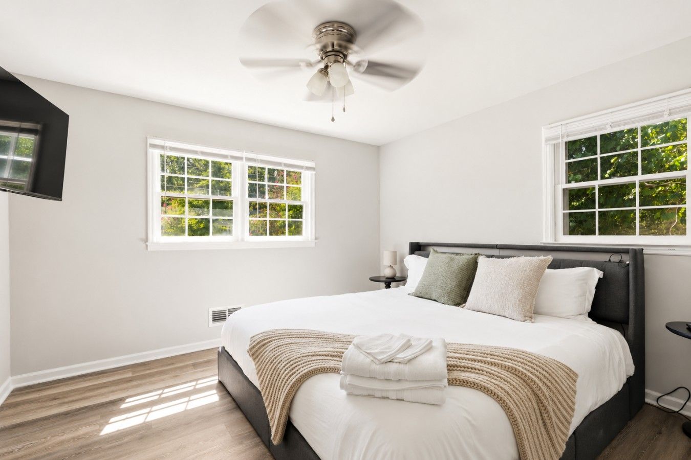 A bedroom with a bed, a ceiling fan, two windows, and a television mounted on the wall.