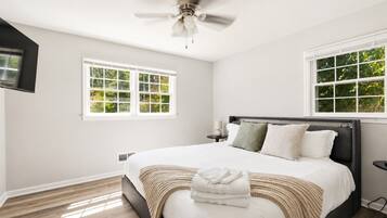 A bedroom with a bed, a ceiling fan, two windows, and a television mounted on the wall.