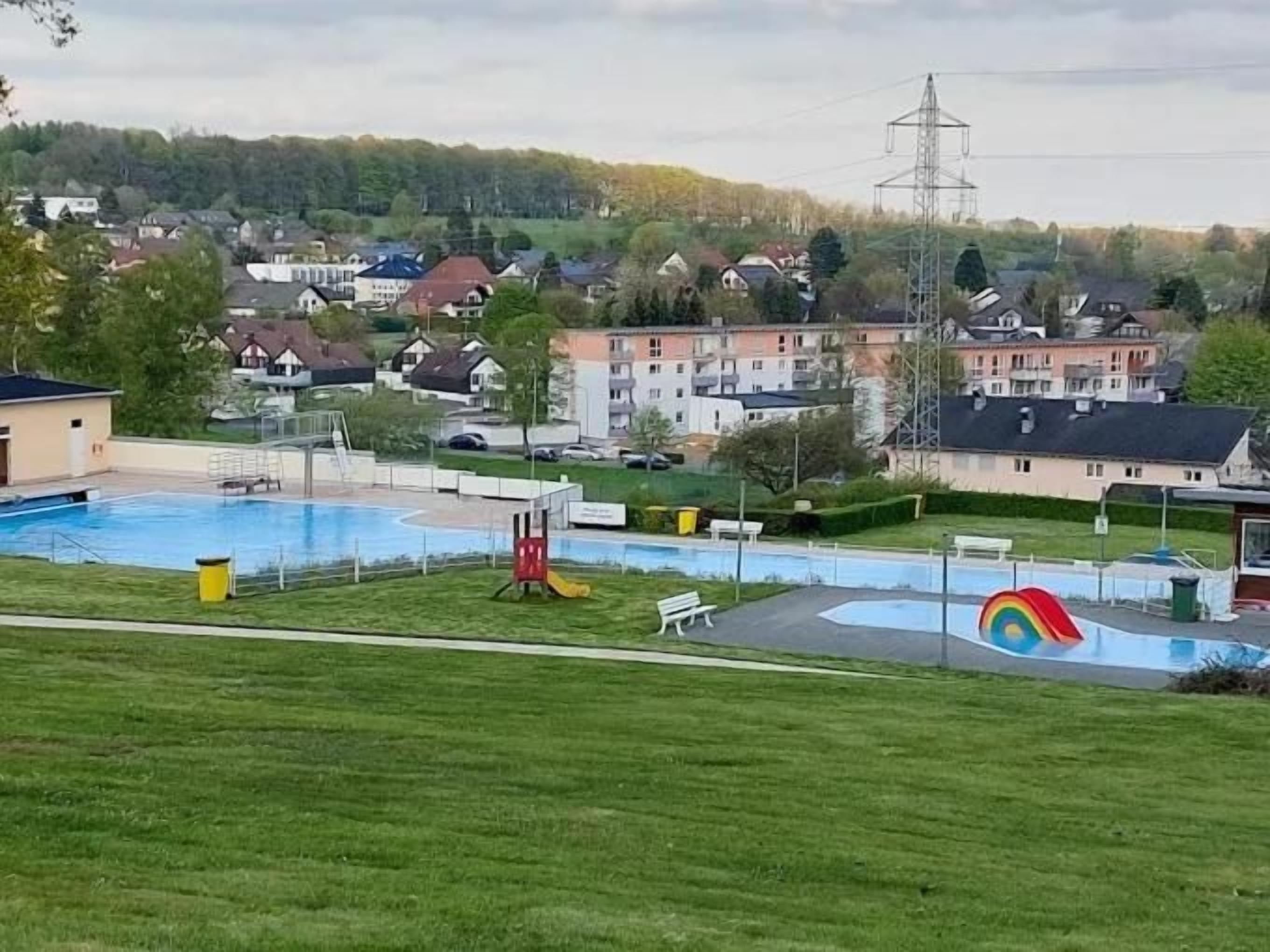 Una piscina con una torre de salvavidas y un mural de arcoíris, rodeada de césped y edificios residenciales al fondo.